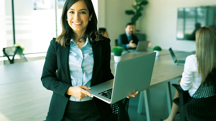 Zu sehen ist eine Businessfrau, die lächelnd einen Laptop hält.
