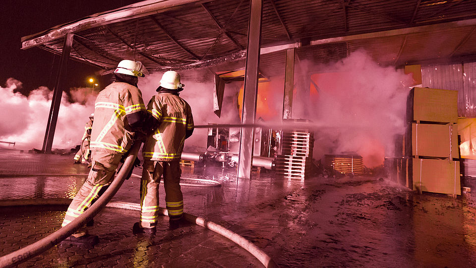 Zu sehen sind zwei Feuerwehrleute beim Löschen eines Lagerhallenbrands.